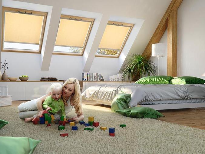Een moeder speelt met haar kind op de slaapkamervloer, op de achtergrond 3 houten dakramen met bijna gesloten rolgordijnen binnenin.